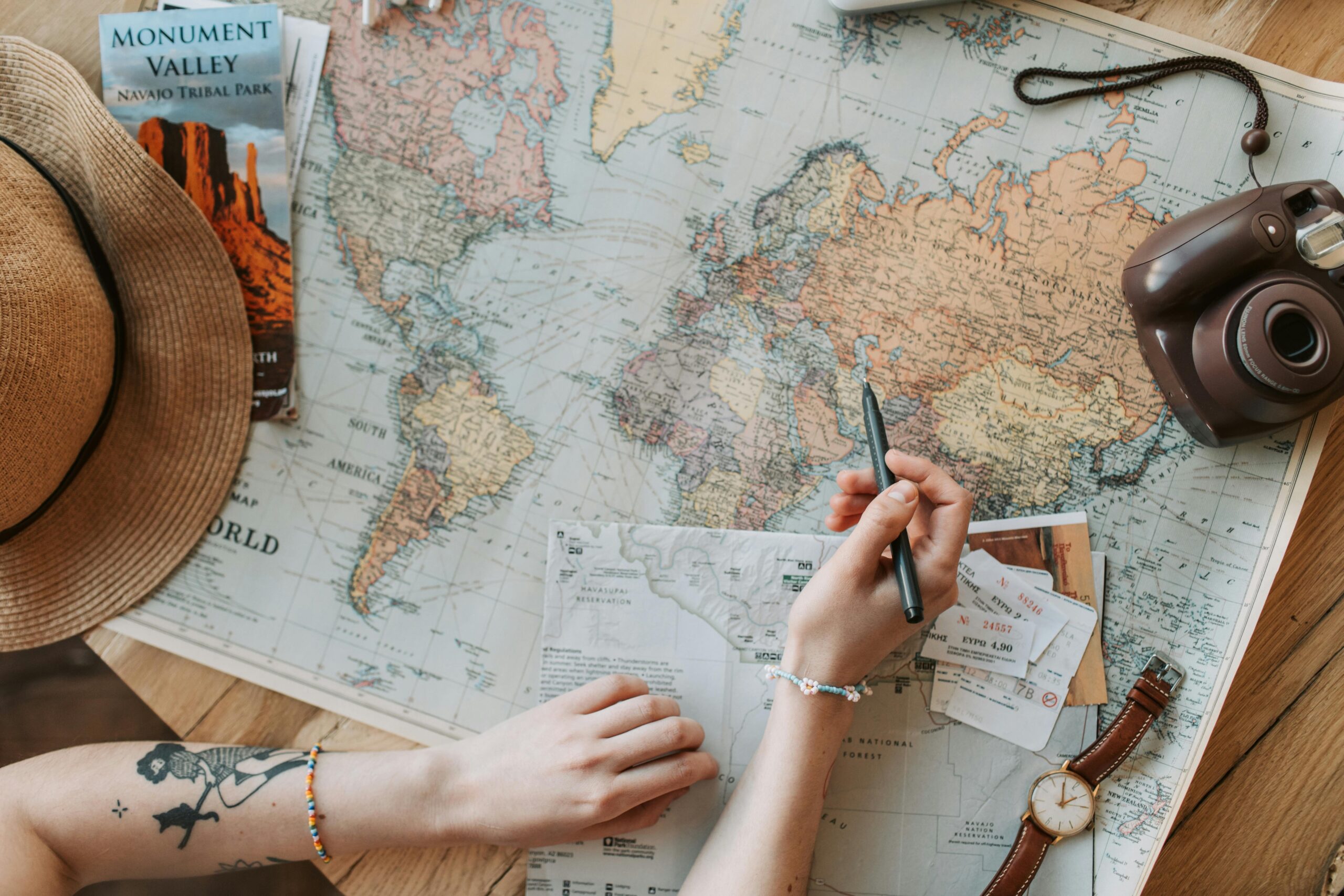 Person planning a journey with a world map, camera, and travel guide on a wooden table.